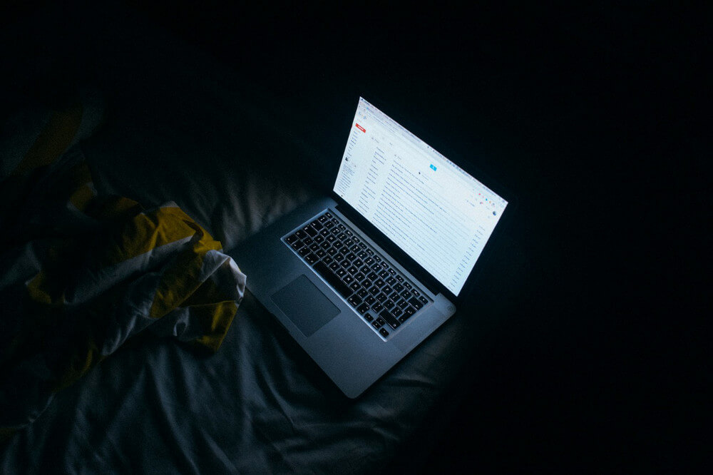 Picture of a laptop, laying on a bed in a dark room, and emitting tons of blue light.